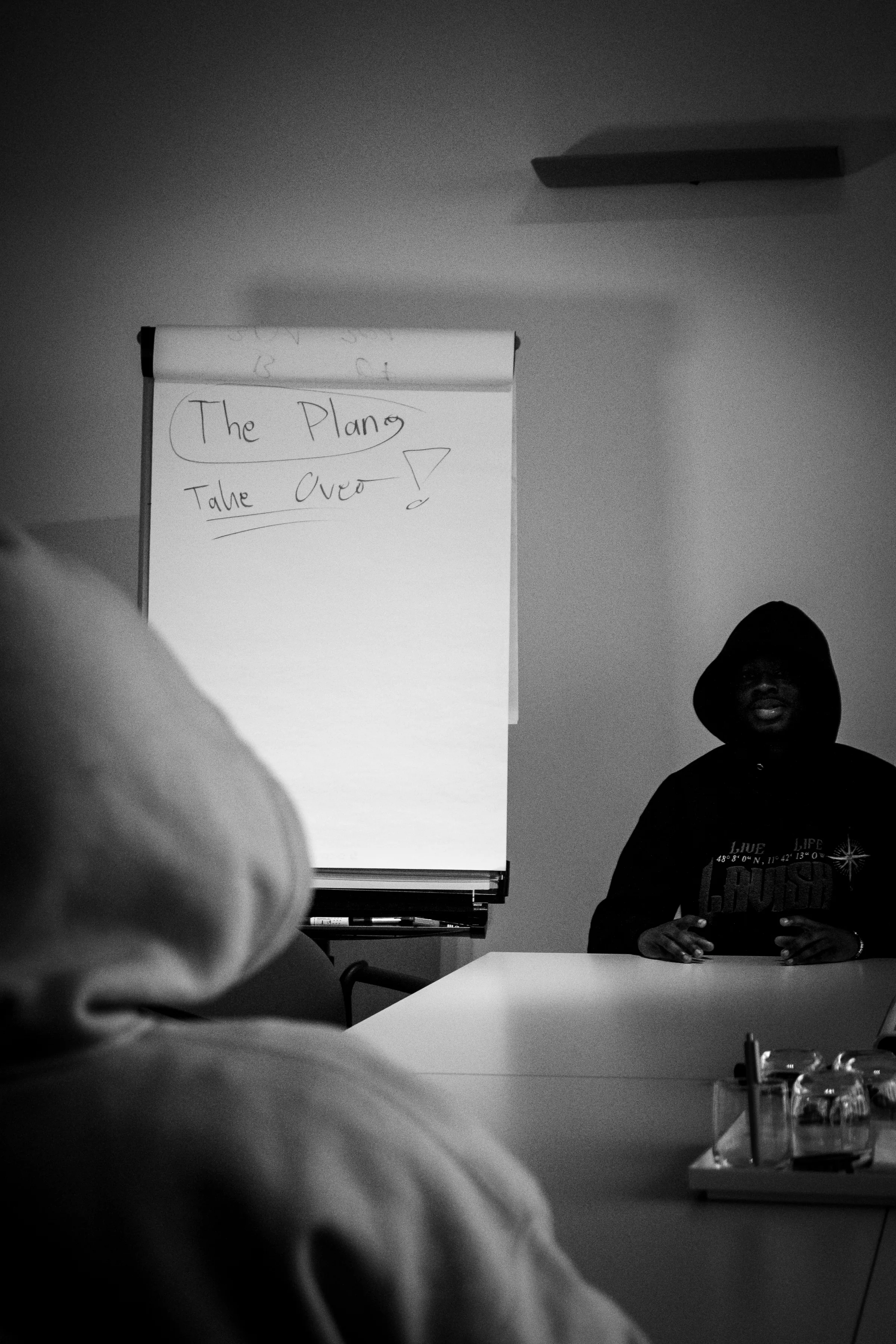 Zwei Personen mit Kapuze sitzen sich an einem Tisch gegenüber, im Hintergrund ein Flipchart – Schwarzweißaufnahme aus der lllavishclo-Serie